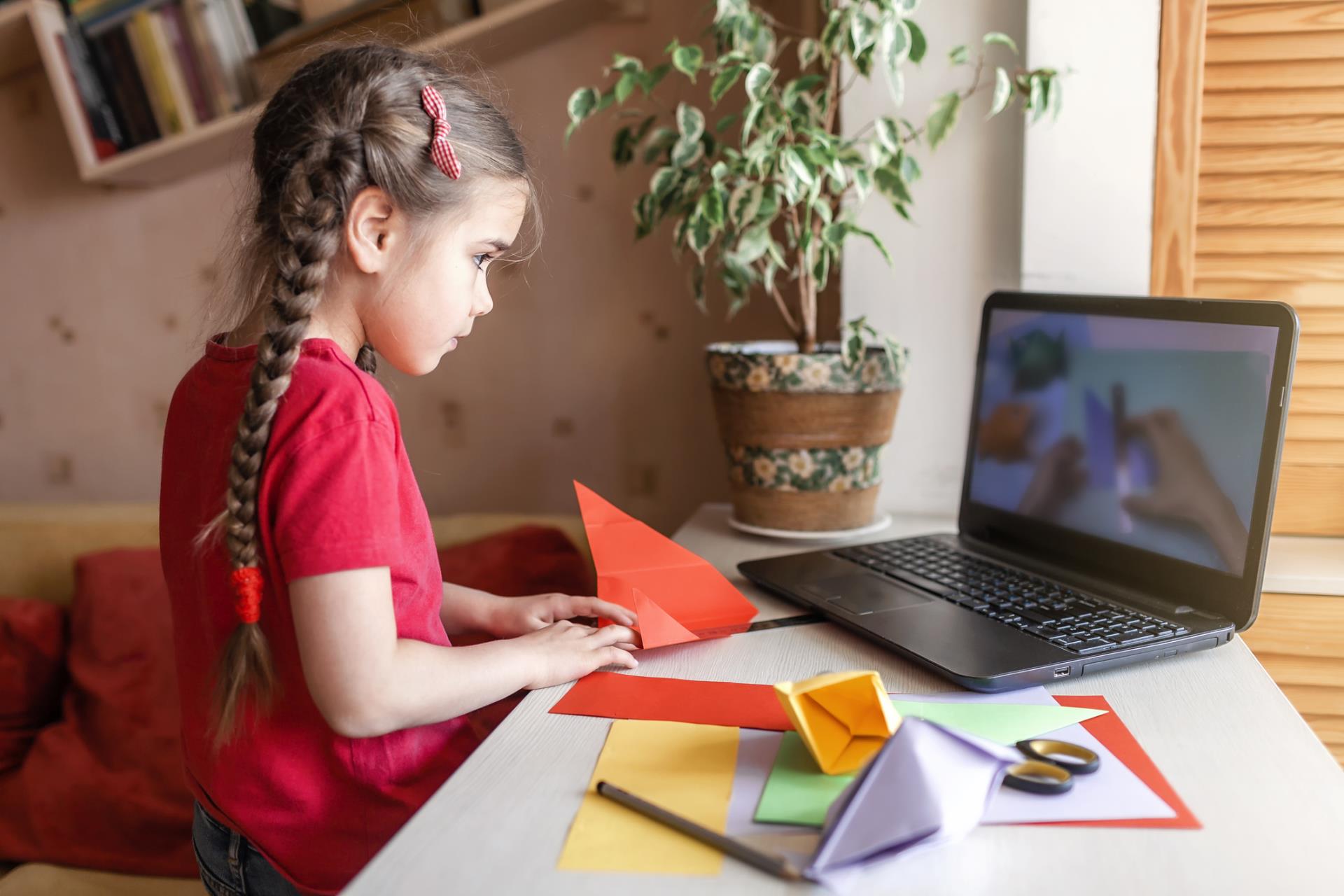 Child completing an origami project while following a lesson on their laptop.