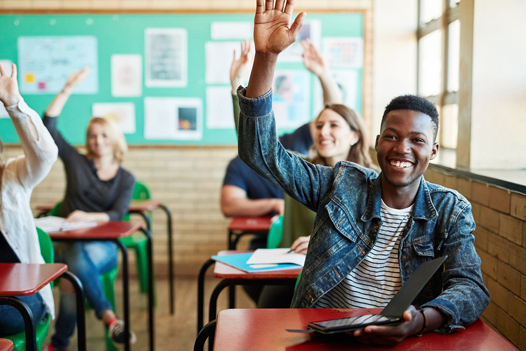 High school student seated at their desk with their hand raised.